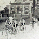 A bunch of cyclists riding in the unpaved, dusty road in front of a villa during Tour de France 1906